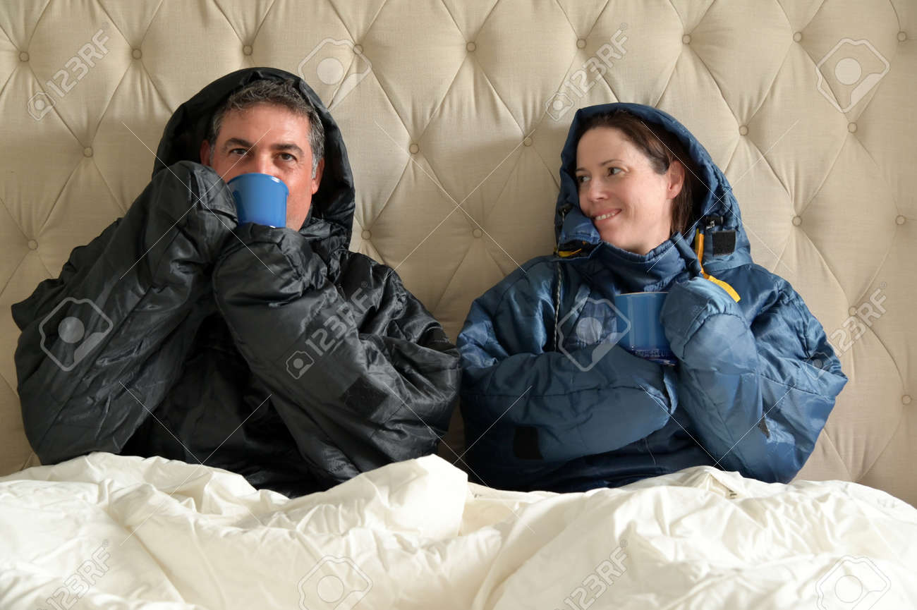 Happy couple sitting side by side wearing a very warm clothing and covered with a blanket, sitting on a double bed, having a hot drink in bedroom on a cold winter day. - 169904523 Happy couple sitting side by side wearing a very warm clothing and covered with a blanket, sitting on a double bed, having a hot drink in bedroom on a cold winter day. - 169904523