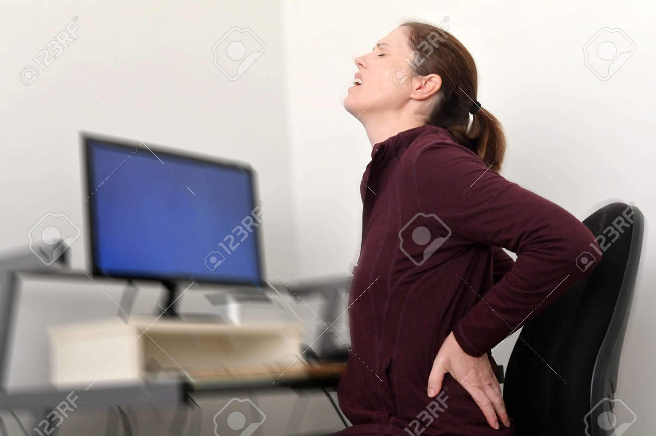 Woman working on a computer at a home office is suffering from lower back pain. - 169904719 Woman working on a computer at a home office is suffering from lower back pain. - 169904719