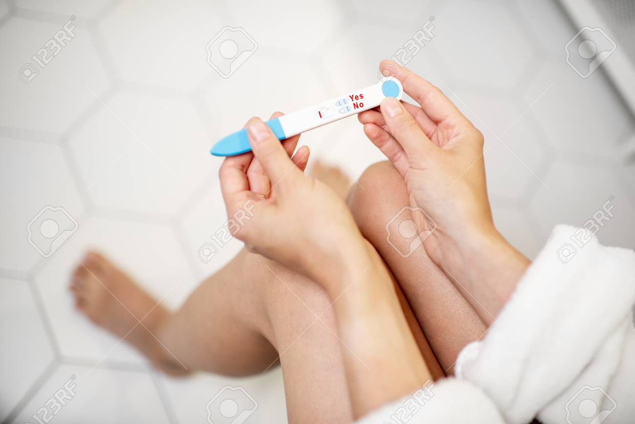 Woman holding pregnancy test stick, sitting on the toilet in the bathroom. Close-up view with no face - 113339055 Woman holding pregnancy test stick, sitting on the toilet in the bathroom. Close-up view with no face - 113339055