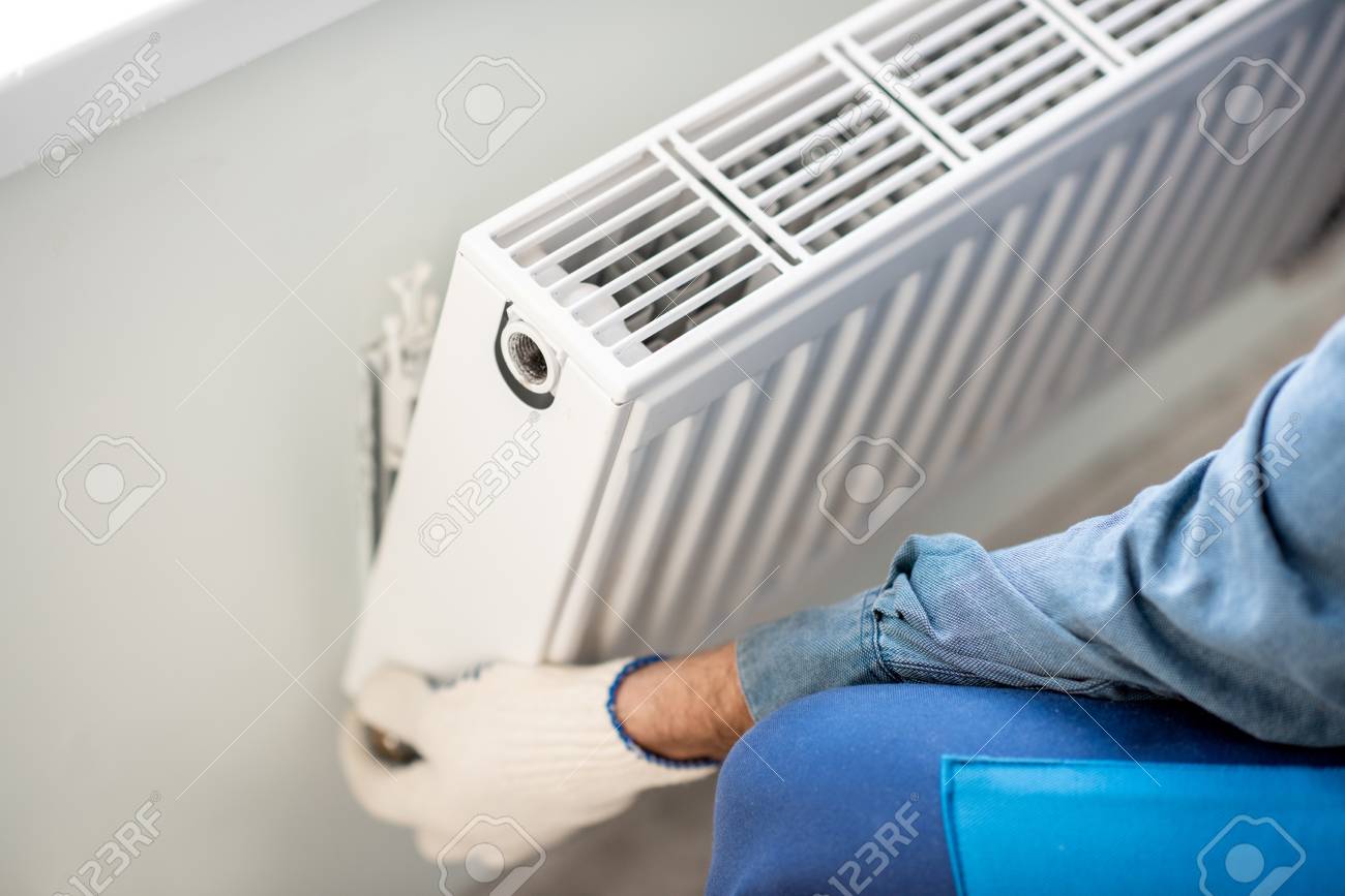 Workman mounting water heating radiator on the white wall indoors, close-up view - 112134302 Workman mounting water heating radiator on the white wall indoors, close-up view - 112134302