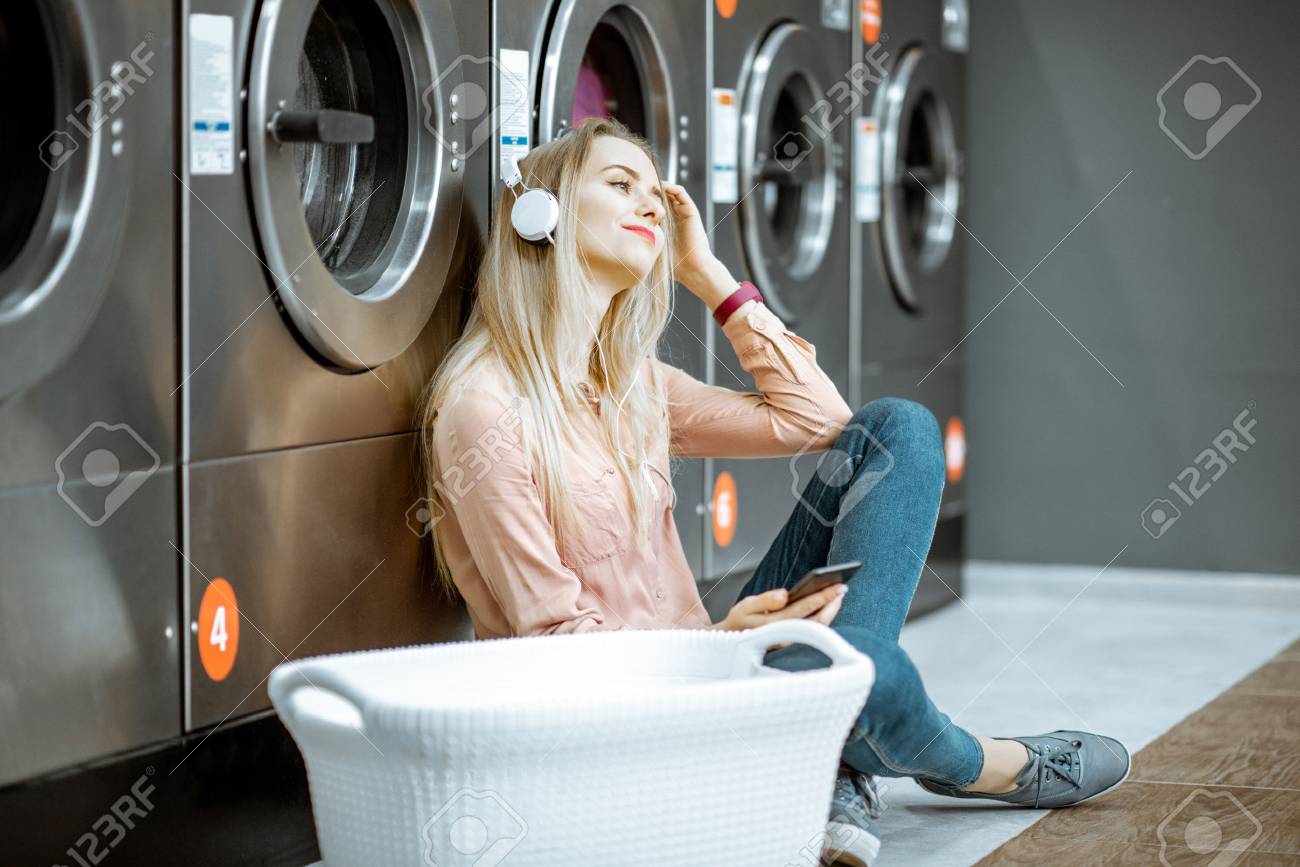 Young woman waiting for the clothes to be washed sitting on the floor and listening to the music at the self-service laundry - 115518762 Young woman waiting for the clothes to be washed sitting on the floor and listening to the music at the self-service laundry - 115518762