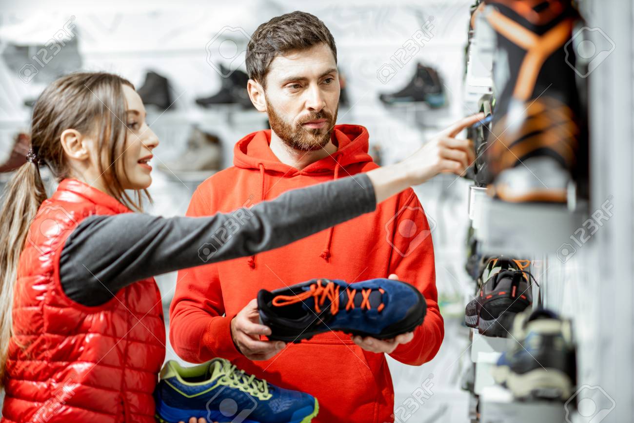 Mam and woman in red sports clothes choosing trail shoes for hiking standing near the showacase of the modern sports shop - 115290187 Mam and woman in red sports clothes choosing trail shoes for hiking standing near the showacase of the modern sports shop - 115290187