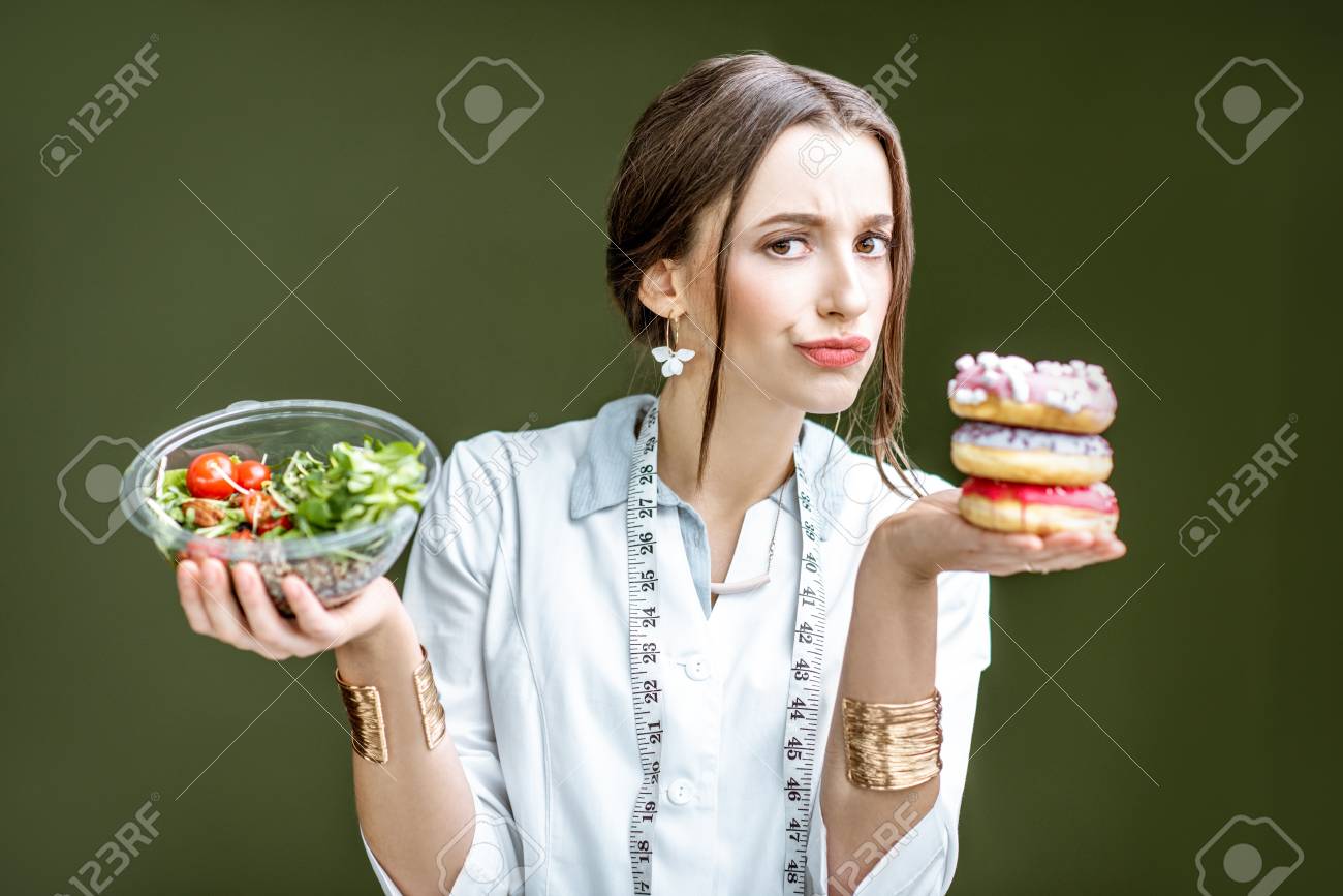 Young woman nutritionist looking on the donuts with sad emotions choosing between salad and unhealthy dessert on the green background - 115514594 Young woman nutritionist looking on the donuts with sad emotions choosing between salad and unhealthy dessert on the green background - 115514594