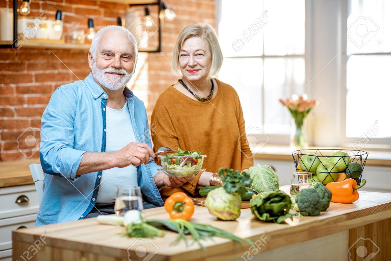 Cheerful senior couple eating salad standing together with healthy food on the kitchen at home. Concept of healthy nutrition in older age - 120618464 Cheerful senior couple eating salad standing together with healthy food on the kitchen at home. Concept of healthy nutrition in older age - 120618464
