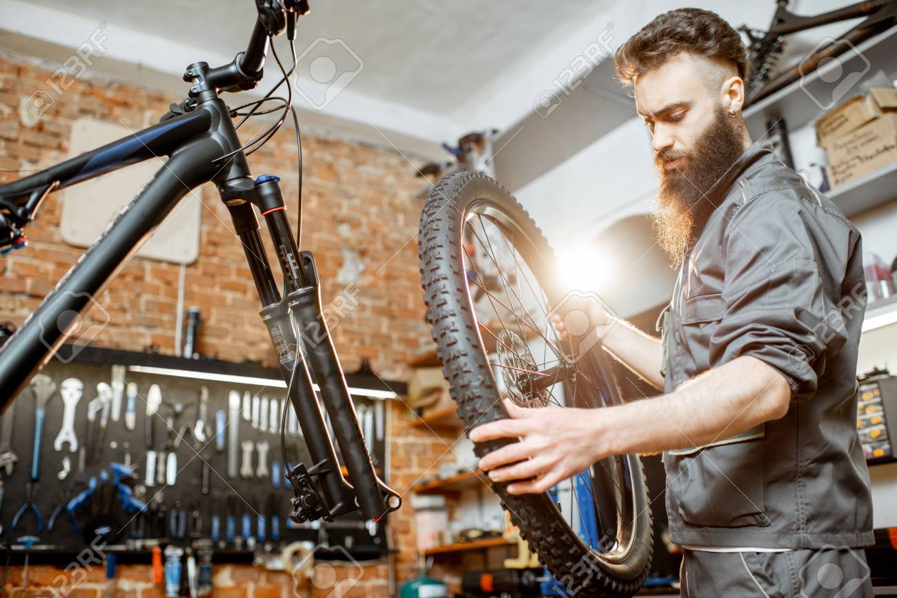 Handsome repairman in workwear serving mountain bicycle, standing with front wheel at the workshop of a bicycle shop - 121320549 Handsome repairman in workwear serving mountain bicycle, standing with front wheel at the workshop of a bicycle shop - 121320549
