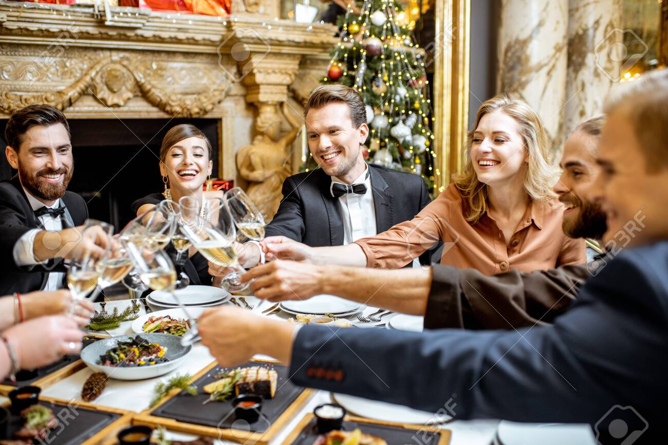 Elegantly dressed group of people having fun, clinking wine glasses during a festive dinner near the fireplace and christmas tree, celebrating New Year holiday at the luxury restaurant - 132844877 Elegantly dressed group of people having fun, clinking wine glasses during a festive dinner near the fireplace and christmas tree, celebrating New Year holiday at the luxury restaurant - 132844877