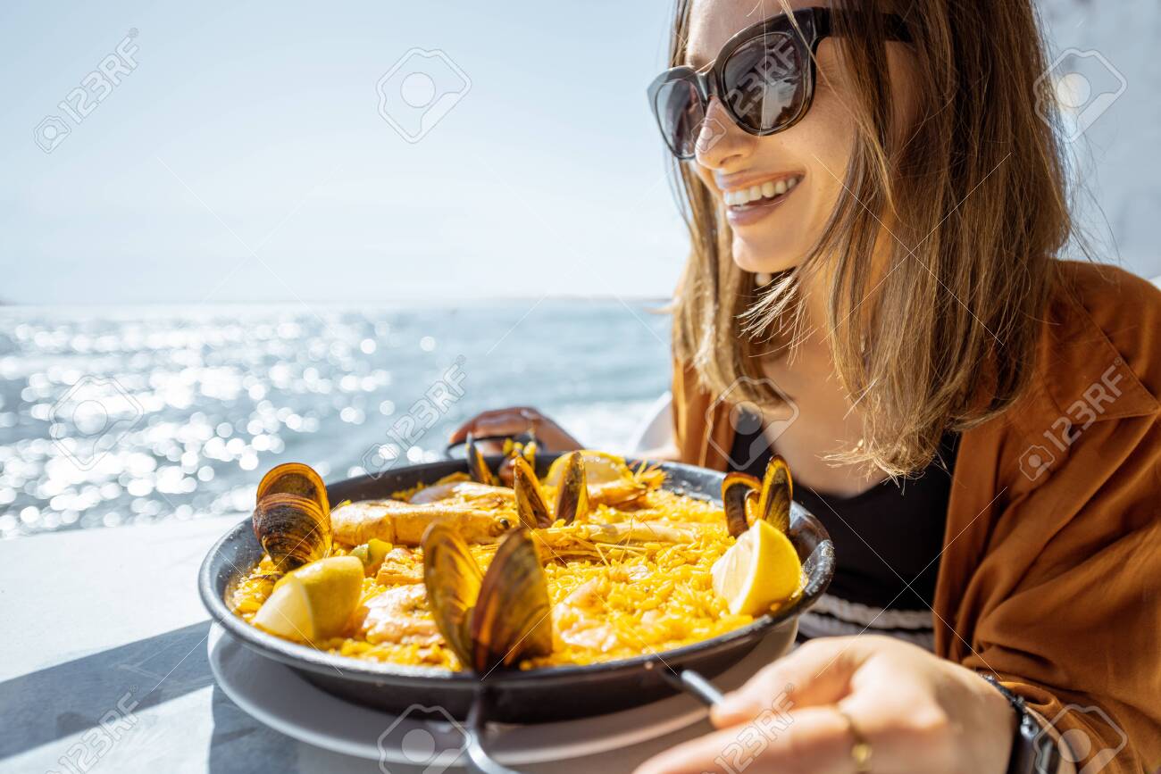 Woman eating paella, traditional spanish dish, while sitting at the restaurant terrace near the ocean. Concept of sea food and good summer vacations - 141789013 Woman eating paella, traditional spanish dish, while sitting at the restaurant terrace near the ocean. Concept of sea food and good summer vacations - 141789013