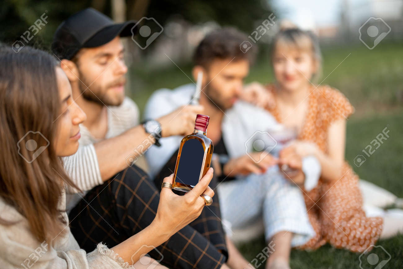Young friends sit together and hug, talk and drink alcohol in a close and friendly atmosphere on a picnic in the evening. Woman holding bottle with blank label to copy paste - 172037487 Young friends sit together and hug, talk and drink alcohol in a close and friendly atmosphere on a picnic in the evening. Woman holding bottle with blank label to copy paste - 172037487