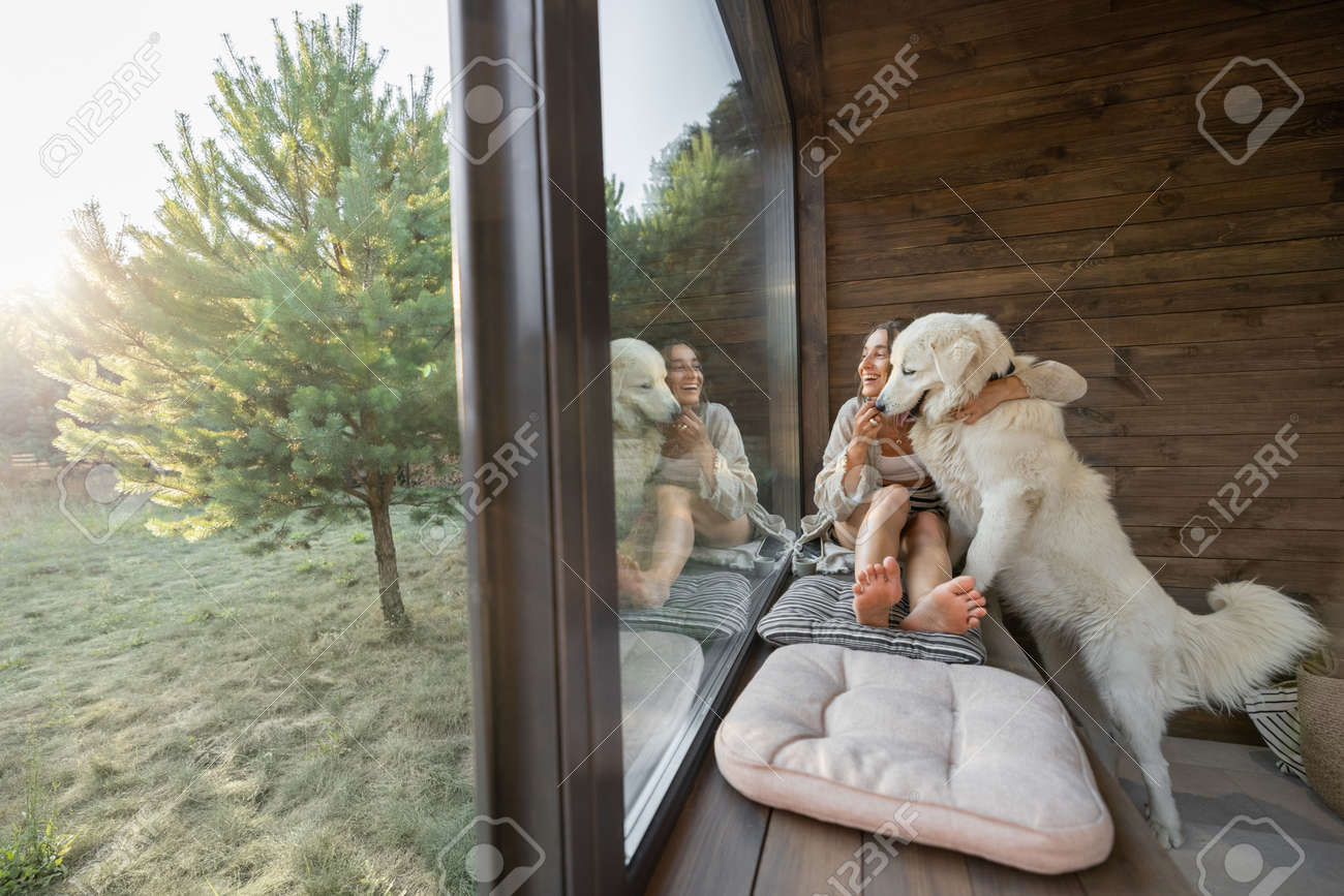 Young woman resting at beautiful country house or hotel, sitting on the window sill with pine forest view and hugs with big white dog. Concept of solitude and recreation on nature with pet - 172224227 Young woman resting at beautiful country house or hotel, sitting on the window sill with pine forest view and hugs with big white dog. Concept of solitude and recreation on nature with pet - 172224227