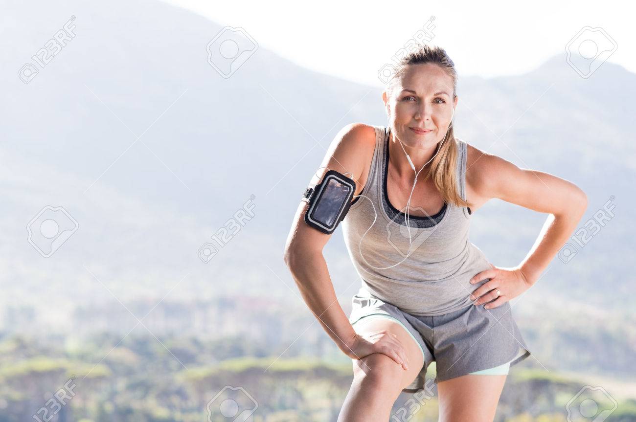 Senior female jogger resting after running in a park. Proud mature woman stretching in the park and looking at camera. Senior active woman completing her daily routine workout. - 56766150 Senior female jogger resting after running in a park. Proud mature woman stretching in the park and looking at camera. Senior active woman completing her daily routine workout. - 56766150