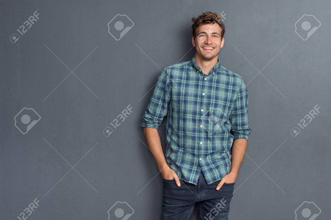 Handsome young man on grey background looking at camera. Portrait of laughing young man with hands in pockets leaning against grey wall. Happy guy smiling. - 61412434 Handsome young man on grey background looking at camera. Portrait of laughing young man with hands in pockets leaning against grey wall. Happy guy smiling. - 61412434