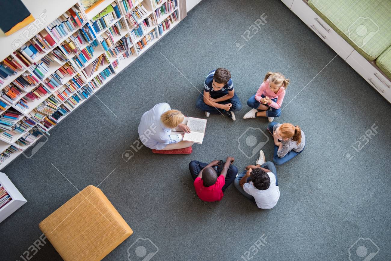 Teacher reading fairy tales to children sitting in a circle at library. Top view of librarian sitting with five multiethnic children on floor. Teacher reading book to cute girls and young boys at school. - 100140682 Teacher reading fairy tales to children sitting in a circle at library. Top view of librarian sitting with five multiethnic children on floor. Teacher reading book to cute girls and young boys at school. - 100140682