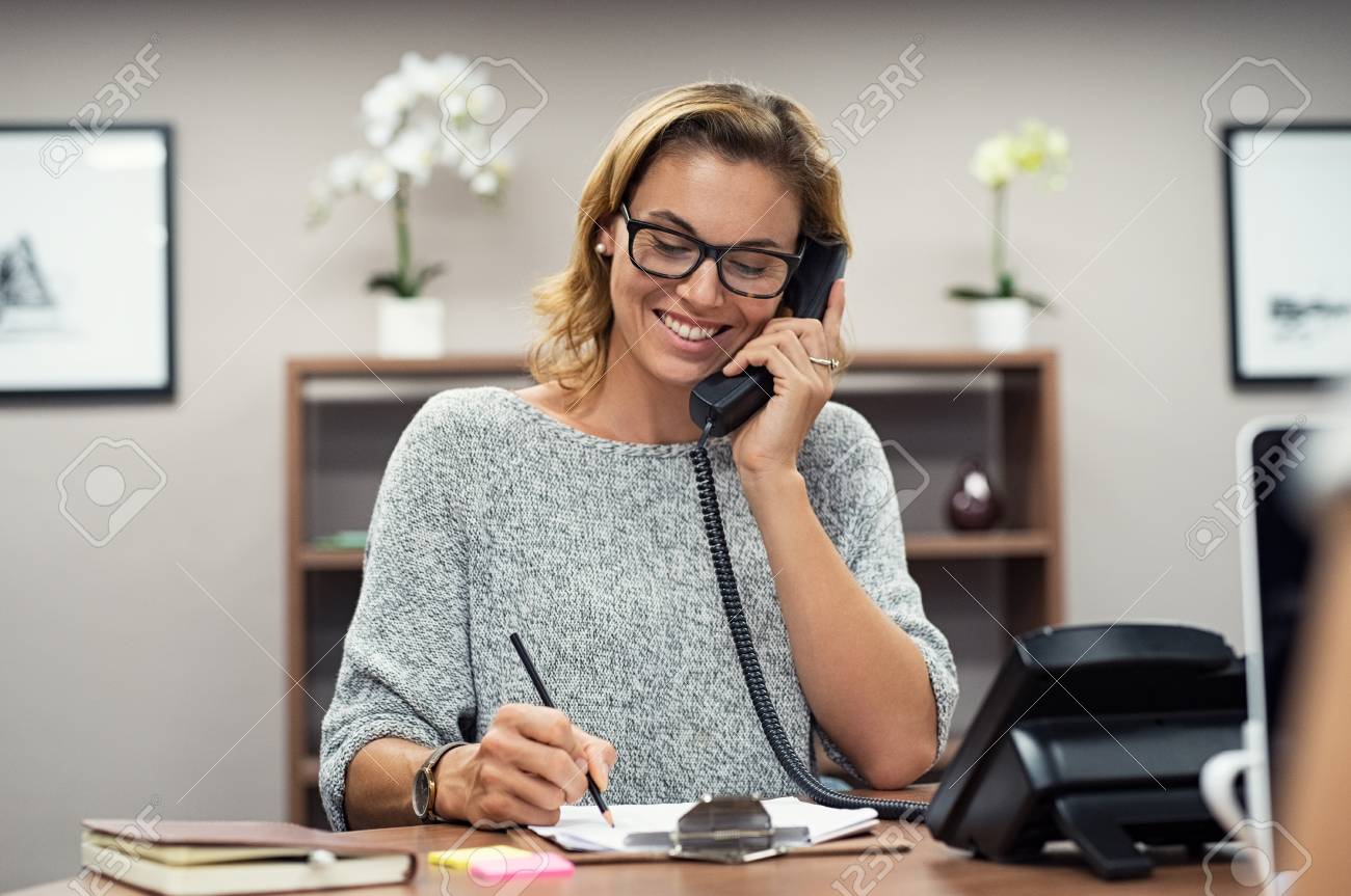 Beautiful mature woman talking on phone at creative office. Happy smiling businesswoman answering telephone at office desk. Casual business woman sitting at her desk making telephone call and taking notes on notebook. - 113992595 Beautiful mature woman talking on phone at creative office. Happy smiling businesswoman answering telephone at office desk. Casual business woman sitting at her desk making telephone call and taking notes on notebook. - 113992595