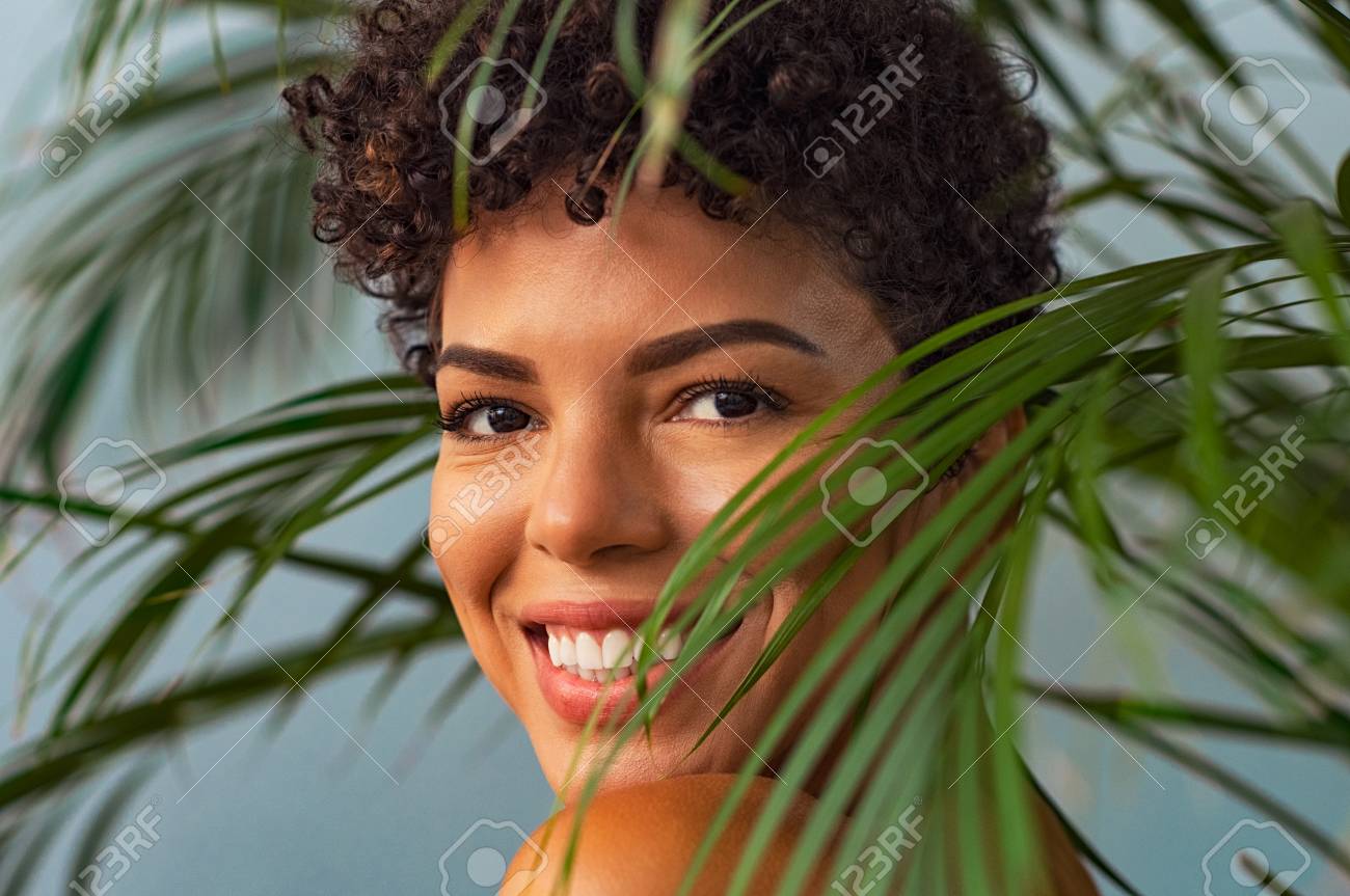 Beautiful Young Woman With Nude Makeup Looking Trought Palm Leaves.  Brazilian Smiling Woman Looking At Camera With Palm Leaves Near Face.  Closeup Face Of Happy African Girl In The Nature. Stock Photo,