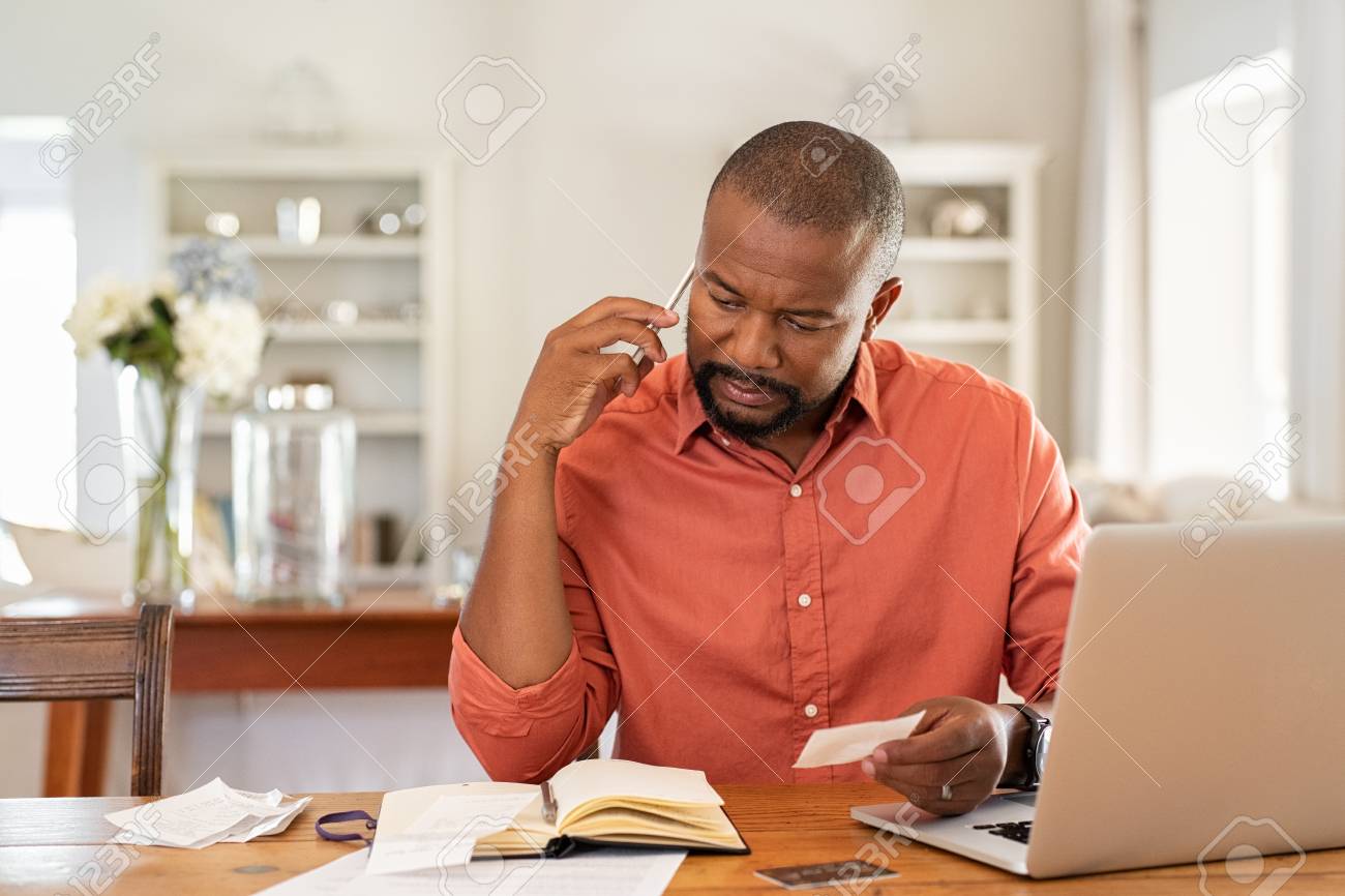 Mature man paying bills with laptop while talking on phone. Thoughtful man at home in conversation over smartphone while checking receipts. Worried african man discussing expenses over phone with bank insurance. - 124983091 Mature man paying bills with laptop while talking on phone. Thoughtful man at home in conversation over smartphone while checking receipts. Worried african man discussing expenses over phone with bank insurance. - 124983091
