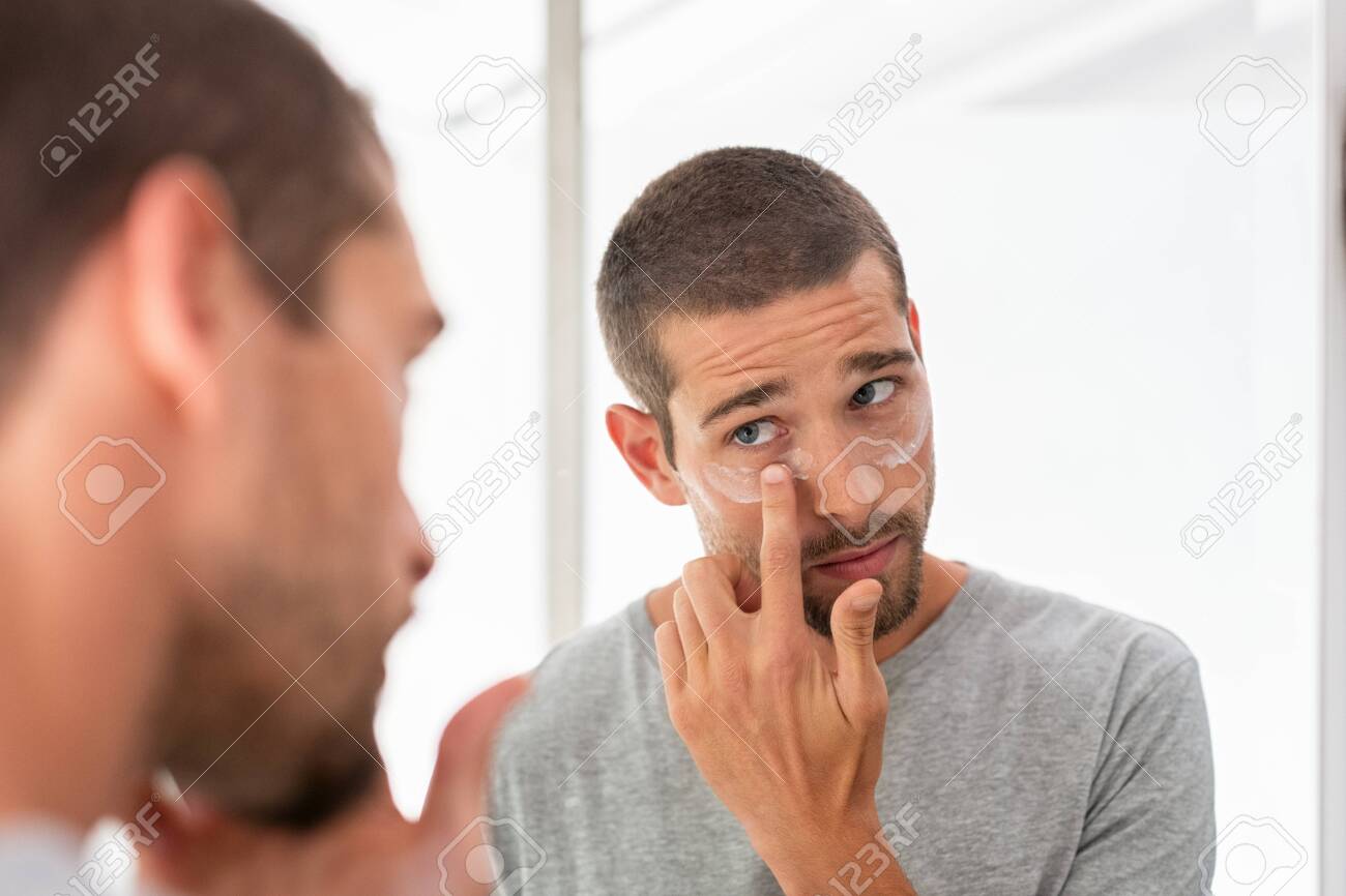 Young man taking care of his undereye wrinkles putting anti aging eye moisturizer. Man applying skincare facial treatment cream on face in the bathroom. Handsome guy applying moisturizer and looking at himself while standing in front of the mirror. - 127284792 Young man taking care of his undereye wrinkles putting anti aging eye moisturizer. Man applying skincare facial treatment cream on face in the bathroom. Handsome guy applying moisturizer and looking at himself while standing in front of the mirror. - 127284792