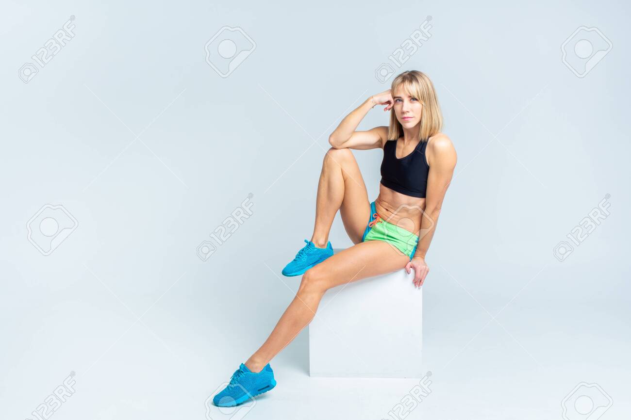 Portrait of beautiful athletic caucasian woman in sport bra and shorts sitting on a white cube on a white studio backgroung. Copyspace - 144199186 Portrait of beautiful athletic caucasian woman in sport bra and shorts sitting on a white cube on a white studio backgroung. Copyspace - 144199186