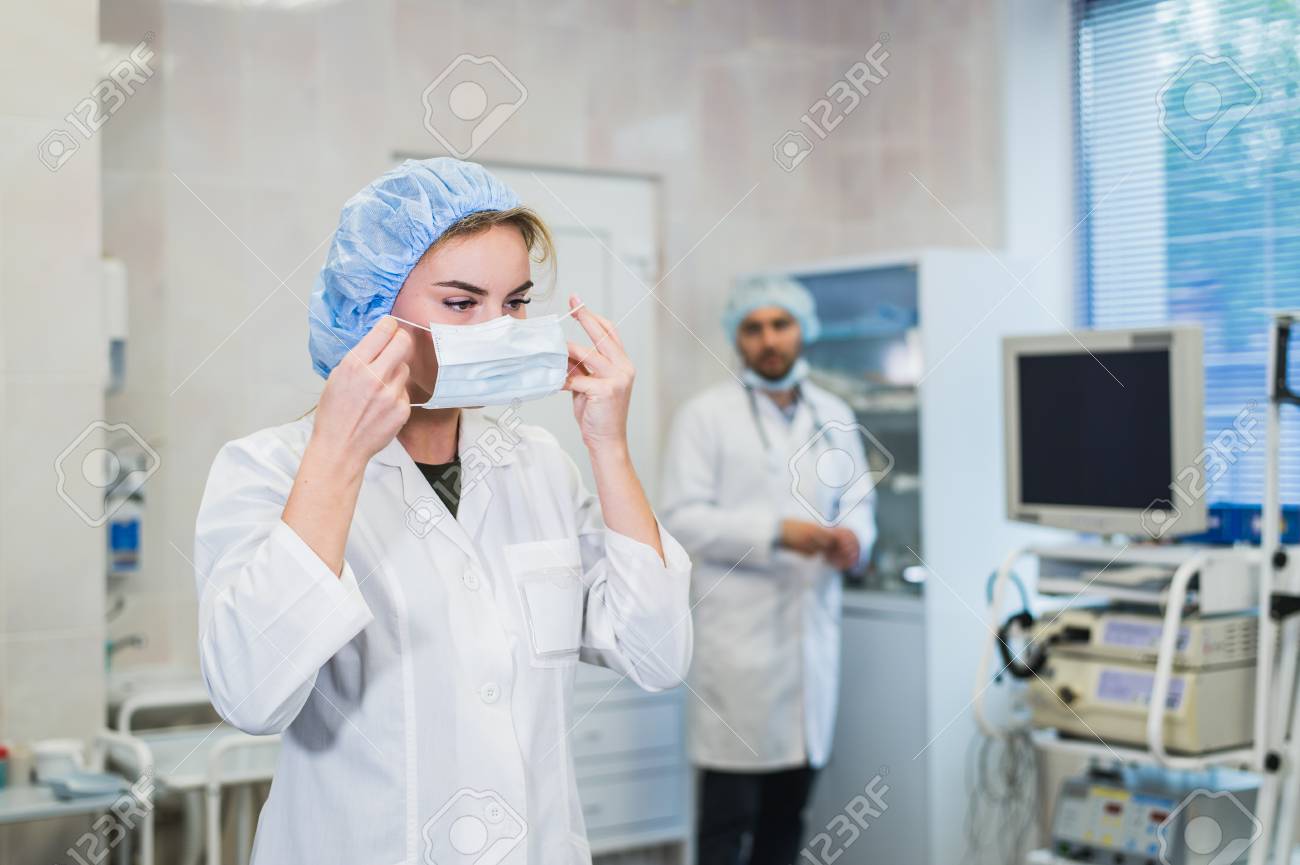 Confident female doctor putting on medical face mask while preparing for operation, her male colleague standing behind her - 83332446 Confident female doctor putting on medical face mask while preparing for operation, her male colleague standing behind her - 83332446