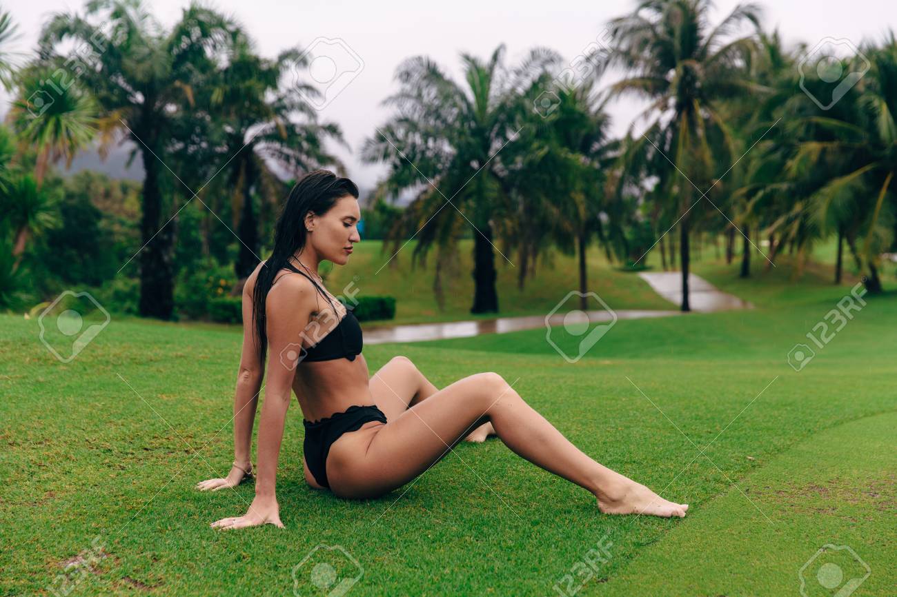 Sexy Young Girl In Bikini Sits On Green Grass, Enjoying Cloudy Wet Weather.  A Model With A Beautiful Body Rests In A Golf Club, Sits On The Grass  Against The Backdrop Of