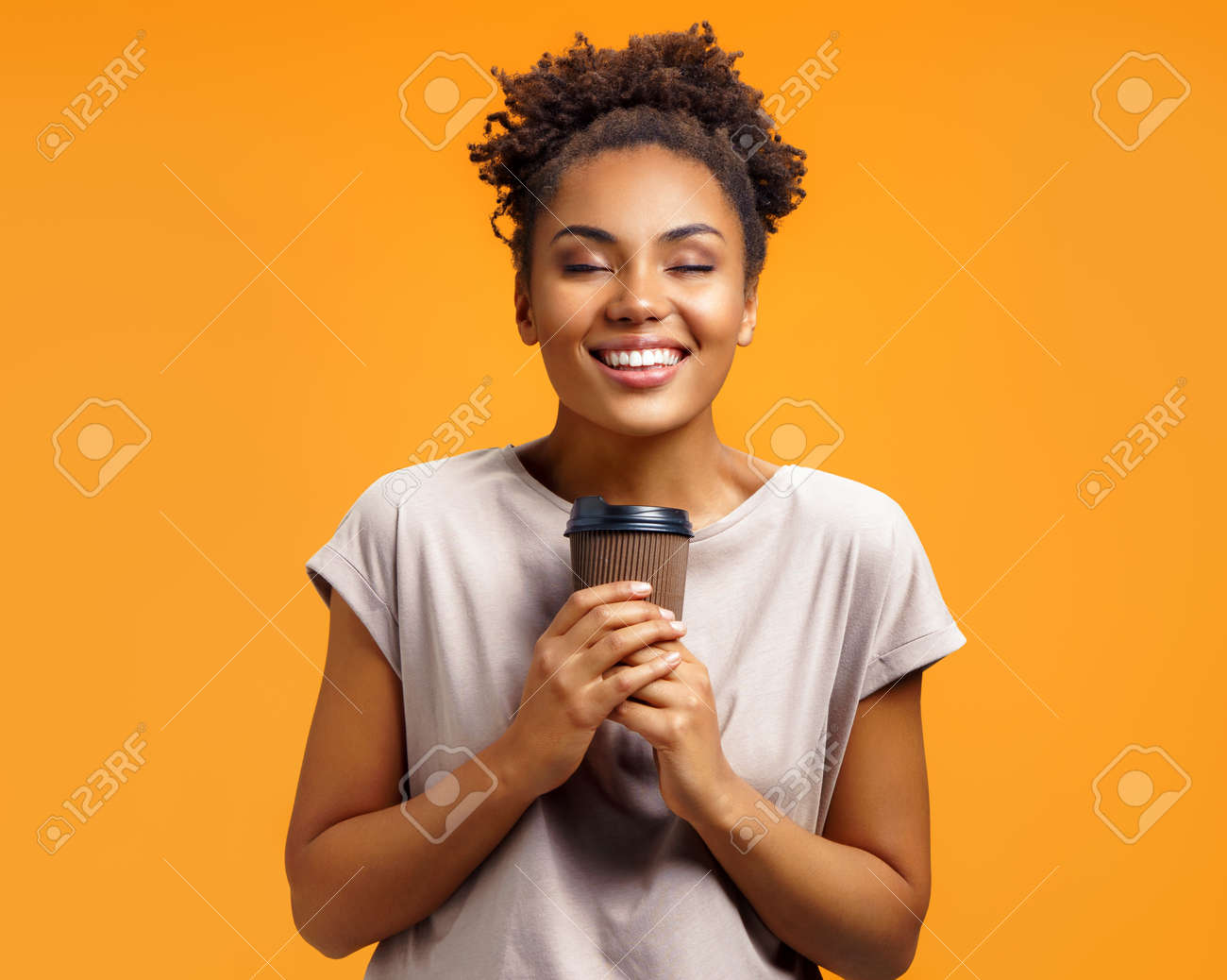 Young girl holds hot takeaway coffee, closes eyes from pleasure. Photo of african american girl wears casual outfit on orange background. Emotions and pleasant feelings concept. - 120344461 Young girl holds hot takeaway coffee, closes eyes from pleasure. Photo of african american girl wears casual outfit on orange background. Emotions and pleasant feelings concept. - 120344461