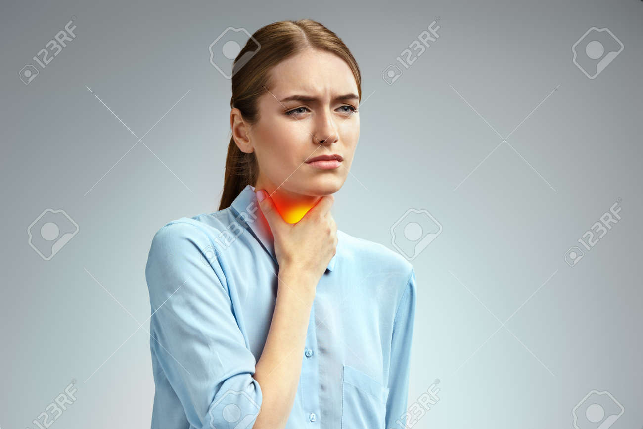 Throat pain. Woman holding her inflamed throat. Photo of american woman in blue shirt on gray background. Medical concept - 123035182 Throat pain. Woman holding her inflamed throat. Photo of american woman in blue shirt on gray background. Medical concept - 123035182
