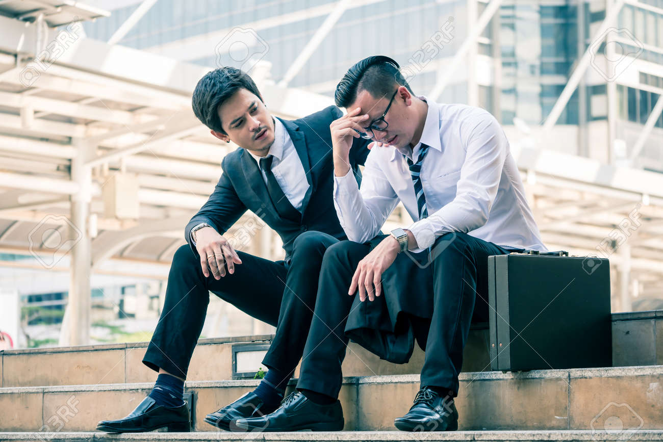 Frustrated Asian young business man sitting outdoor office after he had failed in new project, his colleagues tried to reassure his. - 100743627 Frustrated Asian young business man sitting outdoor office after he had failed in new project, his colleagues tried to reassure his. - 100743627