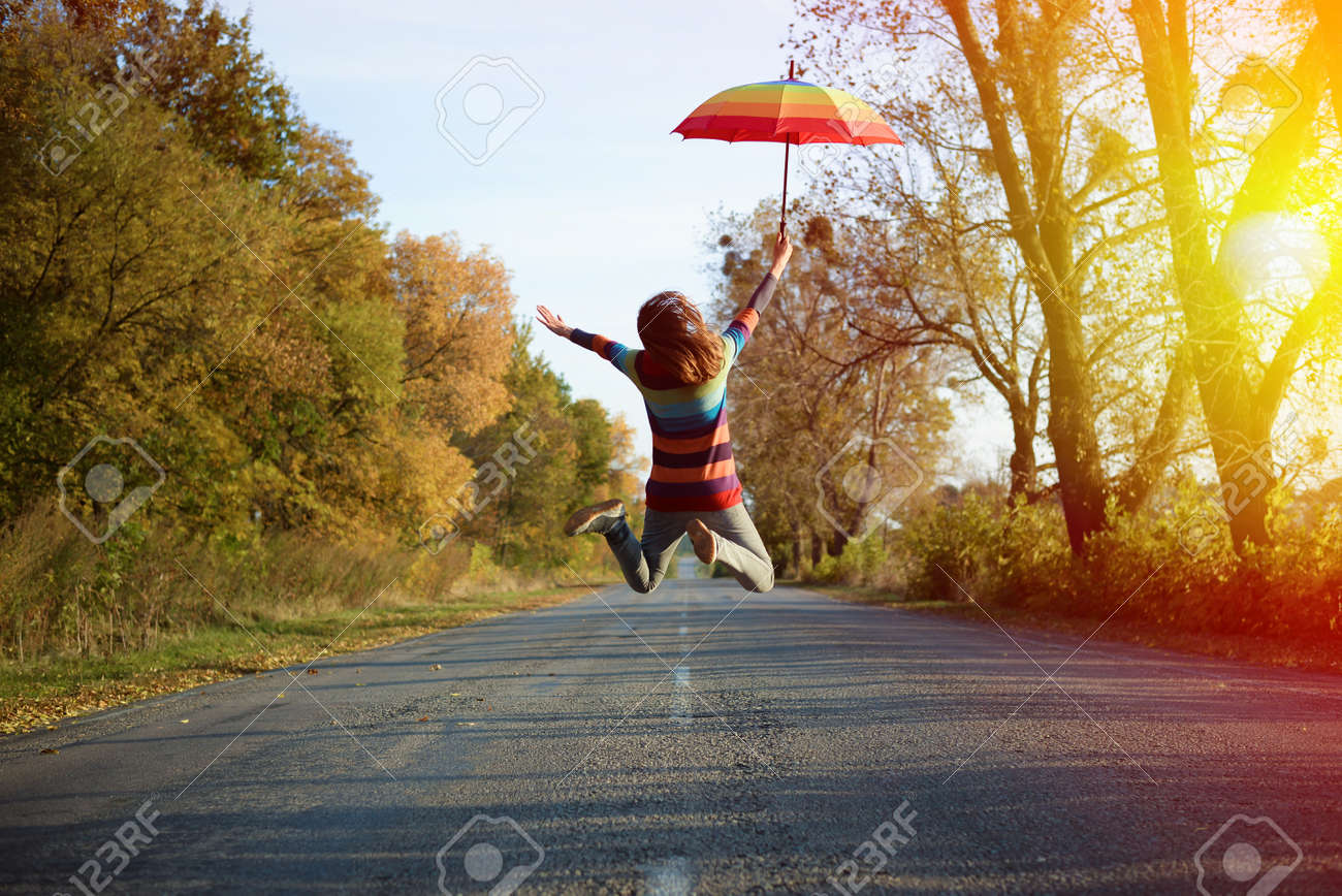Conceptual picture of jumping lady holding umbrella with arms sideways on empty country road in autumn - 45045487 Conceptual picture of jumping lady holding umbrella with arms sideways on empty country road in autumn - 45045487