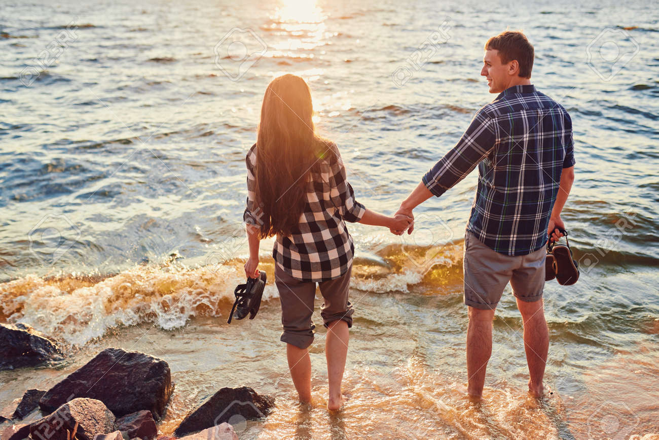 Happy caucasian fashionable couple in love holding hands and walking on coast near river. In background is sunset. - 170687675 Happy caucasian fashionable couple in love holding hands and walking on coast near river. In background is sunset. - 170687675