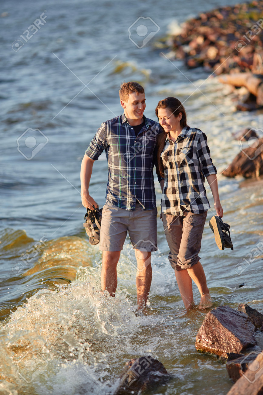 Happy caucasian fashionable couple in love holding hands and walking on coast near river. In background is sunset. - 169020974 Happy caucasian fashionable couple in love holding hands and walking on coast near river. In background is sunset. - 169020974