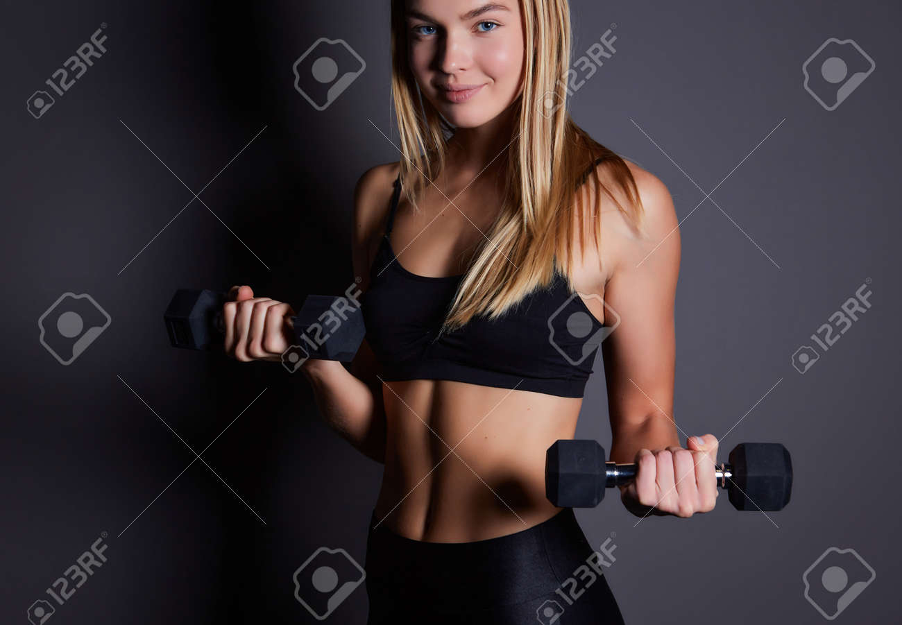 Young fitness blonde woman in the gym near the wall with dumbbells in her hands. - 169021087 Young fitness blonde woman in the gym near the wall with dumbbells in her hands. - 169021087