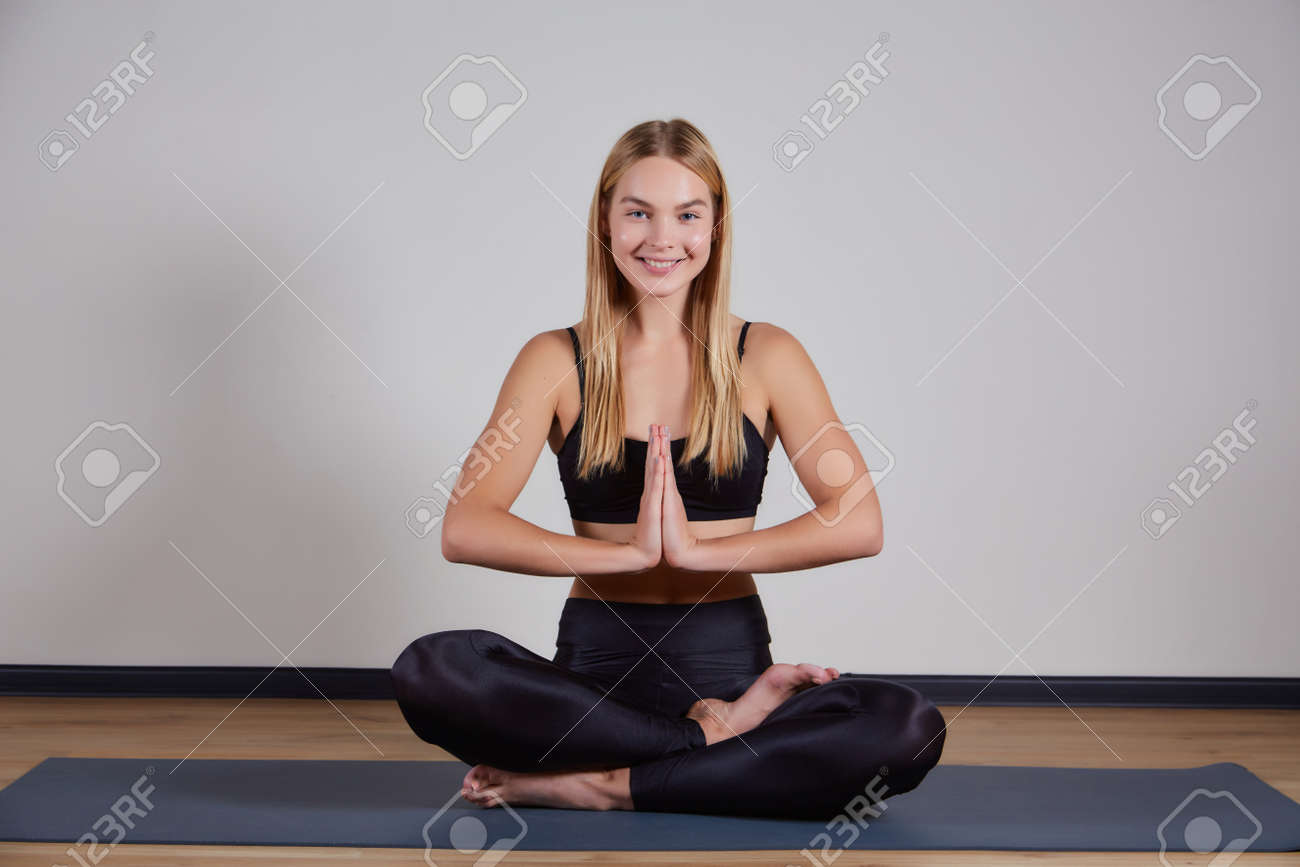 Calm young woman wearing black sportswear practicing yoga, doing Padmasana exercise, sitting in Lotus pose on mat, namaste hands, girl working out at home or in yoga studio with gray walls - 169021091 Calm young woman wearing black sportswear practicing yoga, doing Padmasana exercise, sitting in Lotus pose on mat, namaste hands, girl working out at home or in yoga studio with gray walls - 169021091