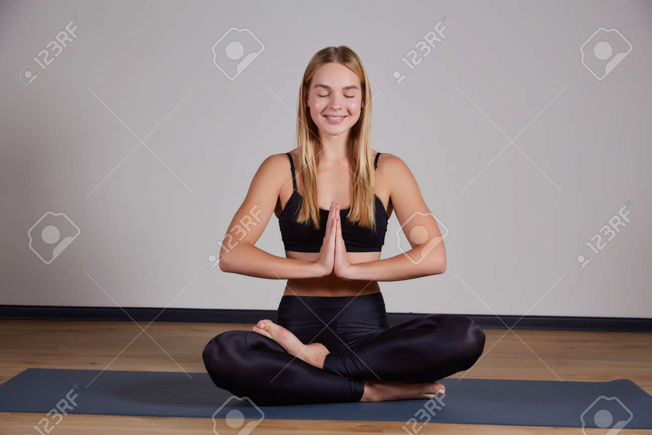 Calm young woman wearing black sportswear practicing yoga, doing Padmasana exercise, sitting in Lotus pose on mat, namaste hands, girl working out at home or in yoga studio with gray walls - 169021093 Calm young woman wearing black sportswear practicing yoga, doing Padmasana exercise, sitting in Lotus pose on mat, namaste hands, girl working out at home or in yoga studio with gray walls - 169021093