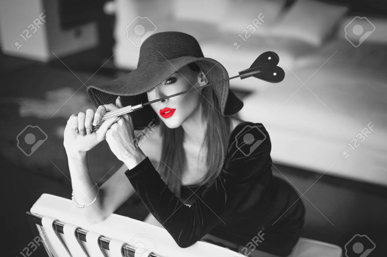 Passionate Dominant Femme Fatale In Hat Holding Whip In Front Of Face,  Kneeling On White Leather Chair In Luxury Flat, Black And White With Red  Lips, Selective Coloring Stock Photo, Picture and, image size:1300x866