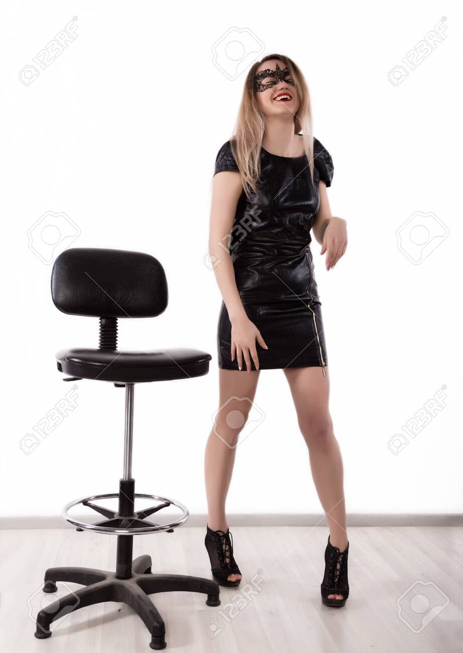 熟女レザーパンツ姿 セクシーな魅惑的な女性は、小さな革の黒いドレスとレースマスクでハイチェアの近くで踊っています。白い背景の女の子の写真素材・画像素材 Image 123016931