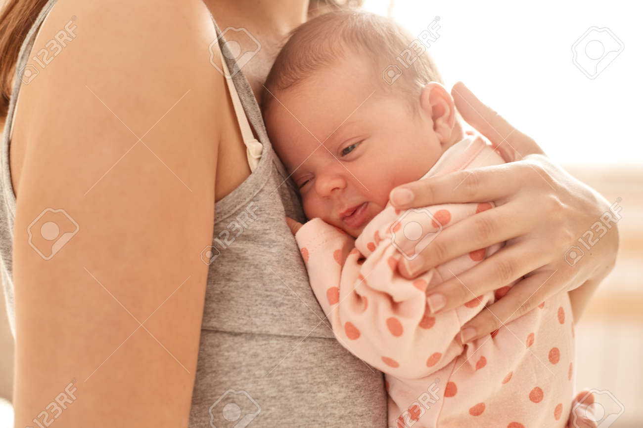Mother holding newborn baby girl on light background, faceless female dresses casual sleeveless t shirt hugging her infant and express love, tiny baby with mommy. - 166345257 Mother holding newborn baby girl on light background, faceless female dresses casual sleeveless t shirt hugging her infant and express love, tiny baby with mommy. - 166345257