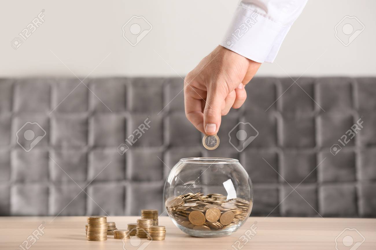 Man putting coins into glass jar on table. Savings concept - 114662646 Man putting coins into glass jar on table. Savings concept - 114662646