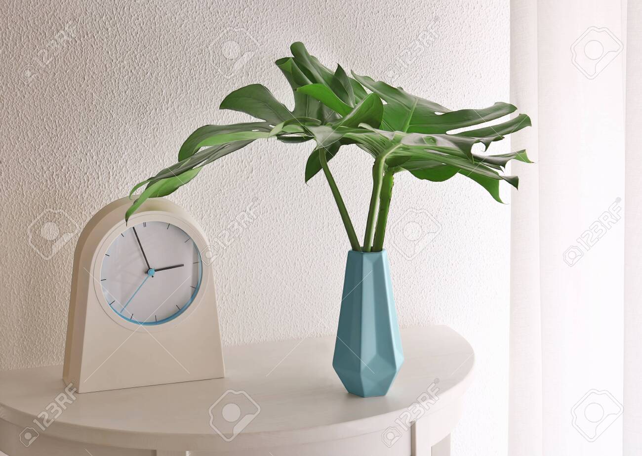 Tropical leaves in vase with clock on table near light wall indoors - 118456187 Tropical leaves in vase with clock on table near light wall indoors - 118456187