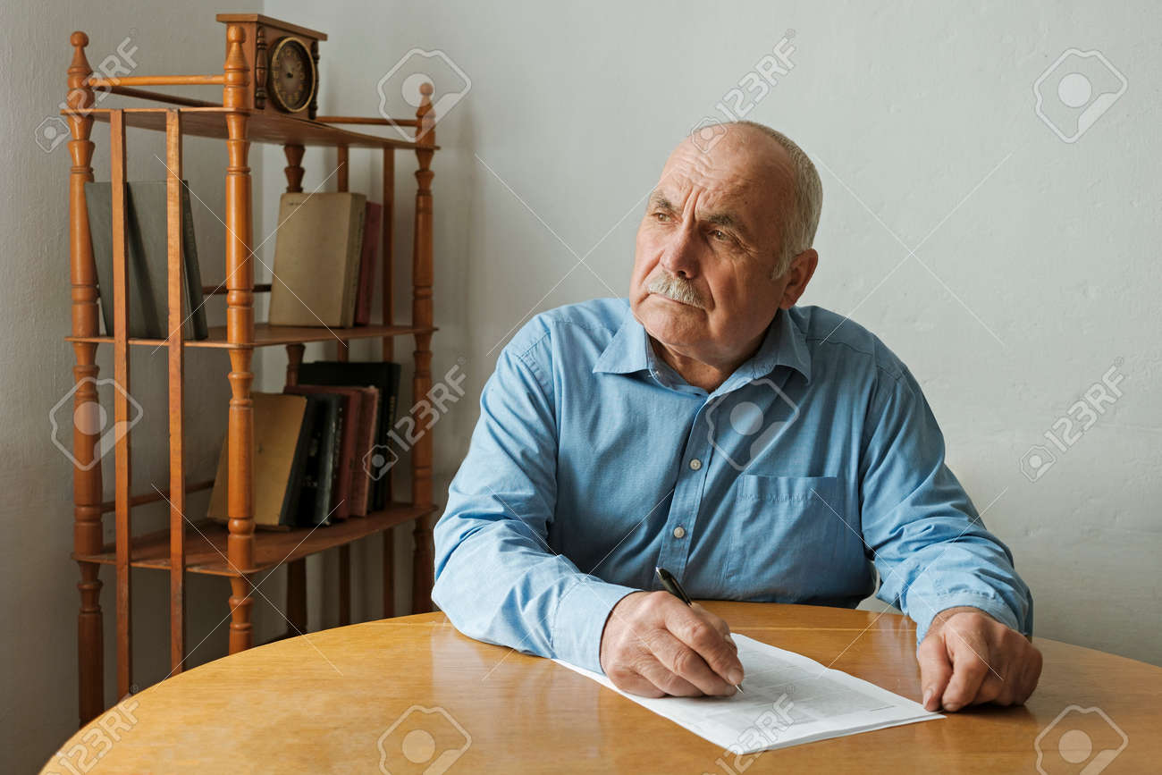 Old Man Writing Notes Or Deciding Whether To Sign A Paper Document On The Table In Front Of Him As He Looks To The Side With A Thoughtful Engrossed Expression Stock Photo