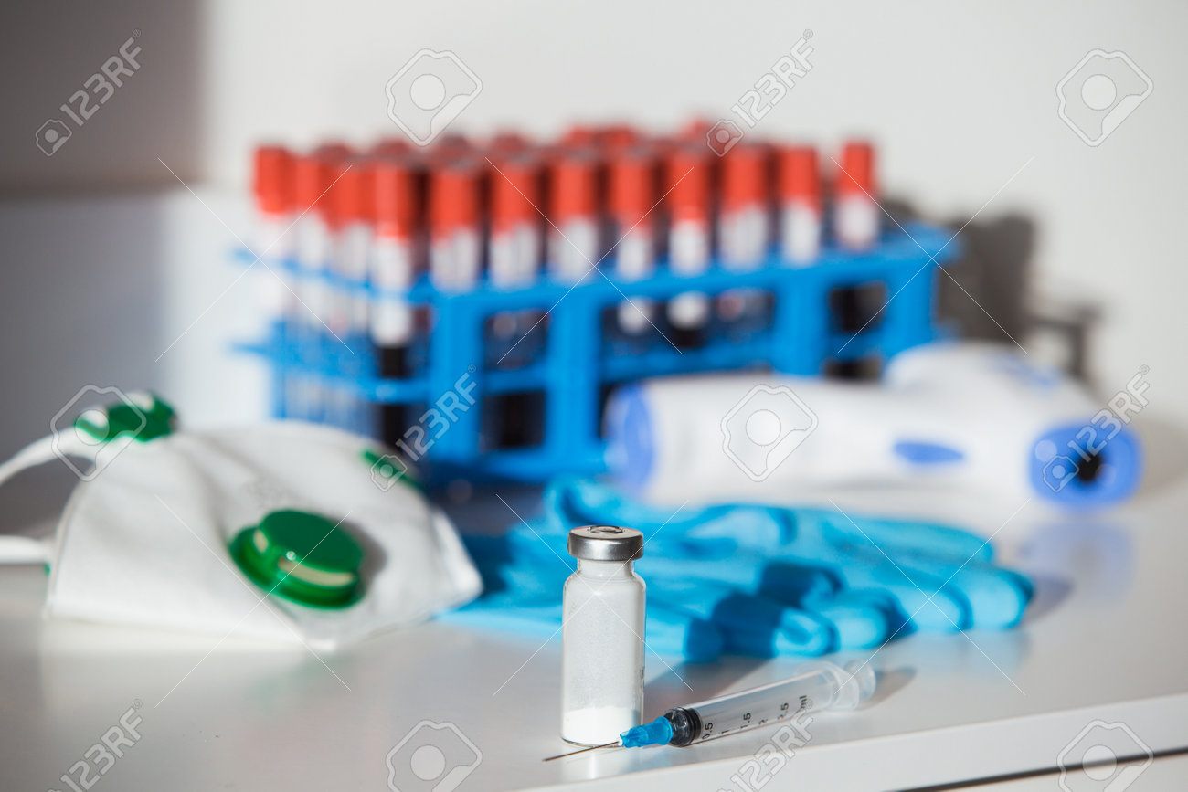 Close up of syringe with Covid-19 vaccine on a white table with protective mask and gloves and blood tube rack. - 160752447 Close up of syringe with Covid-19 vaccine on a white table with protective mask and gloves and blood tube rack. - 160752447