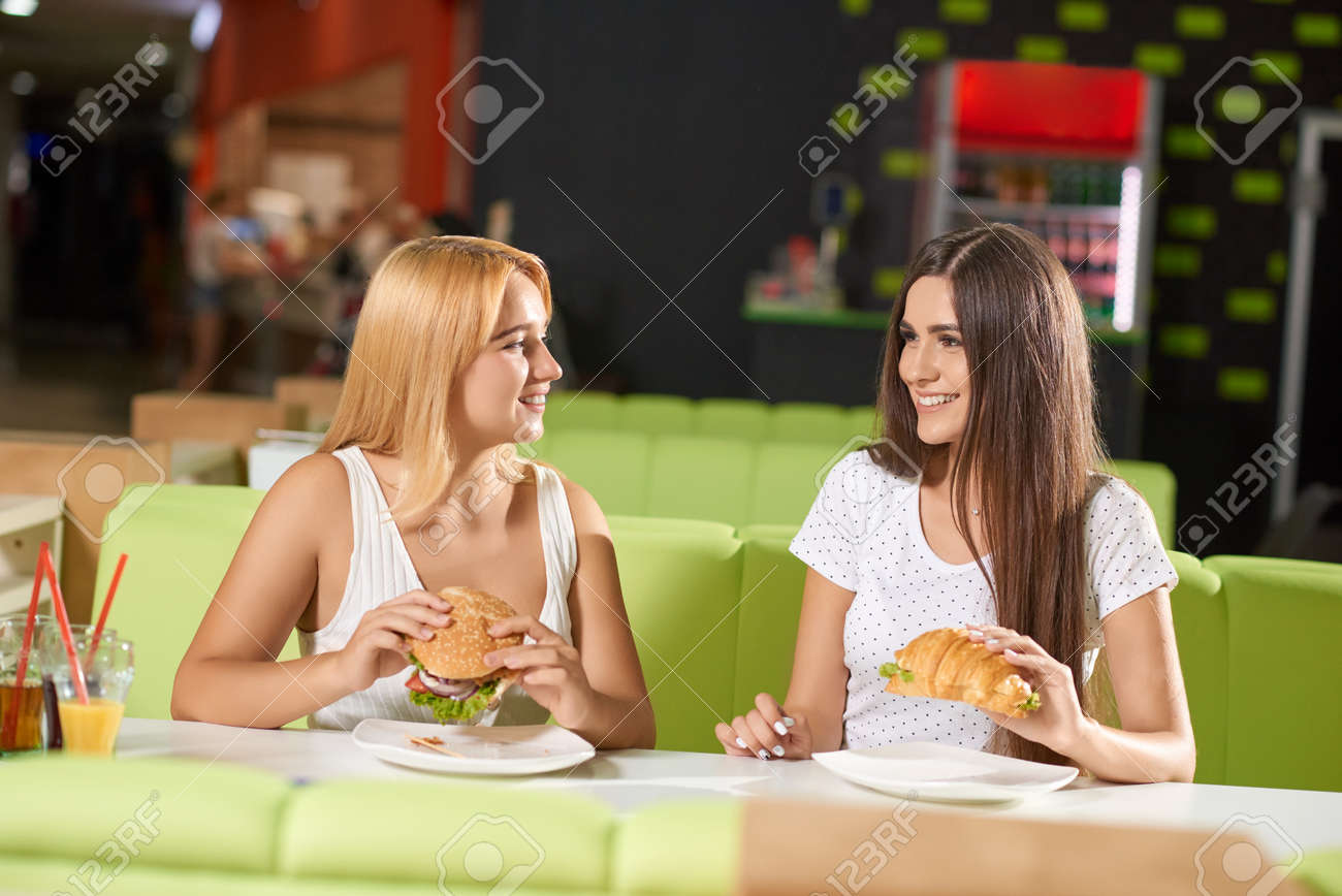 Front View Of Two Attractive Girls Eating Delicious Burger And Croissant In  Cafe. Brunette And Blonde Sitting At Table, Looking At Each Other, Talking  And Laughing. Concept Of Meeting. Stock Photo, Picture
