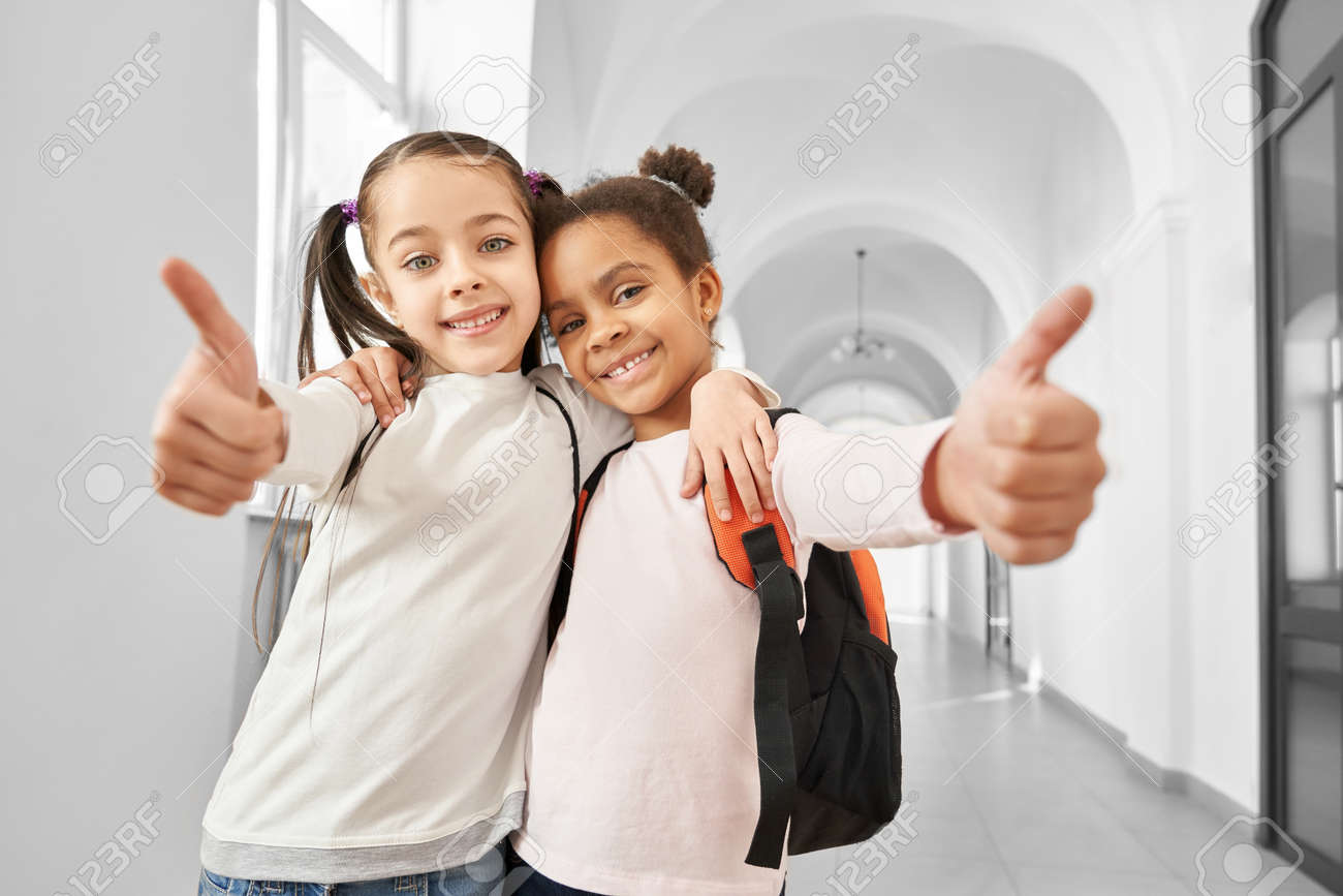 女子小学生　笑顔 かわいい、ポジティブな、チアフールの女子生徒が抱きしめて親指を見せる。幸せな友人は、小学生の長い光の廊下に立って、笑顔で、カメラを見て。の写真素材・画像素材  Image 131828858