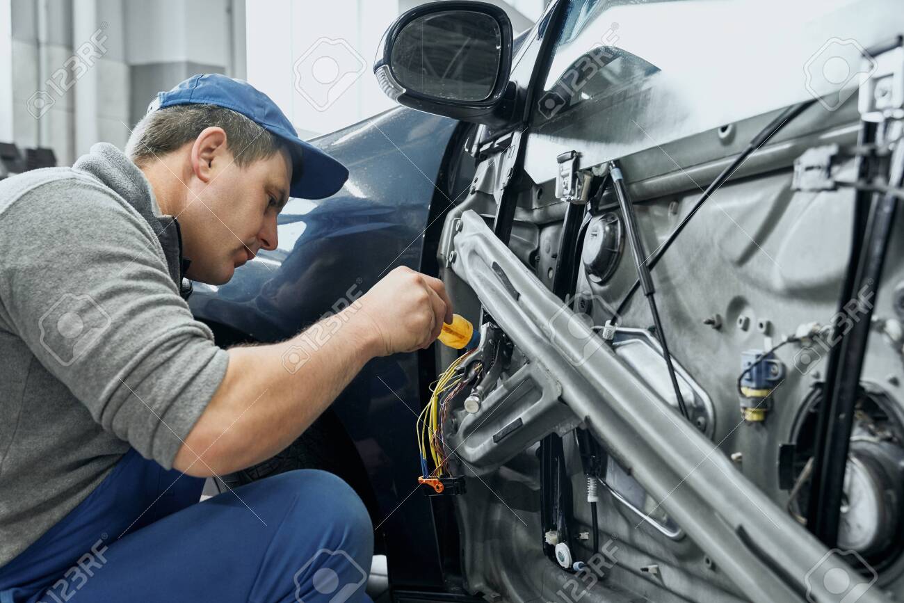 Side view of auto mechanic in blue uniform and cap repairing electrical wires in door of old car. Middle aged man changing damaged details to new ones in car service station - 133752810 Side view of auto mechanic in blue uniform and cap repairing electrical wires in door of old car. Middle aged man changing damaged details to new ones in car service station - 133752810