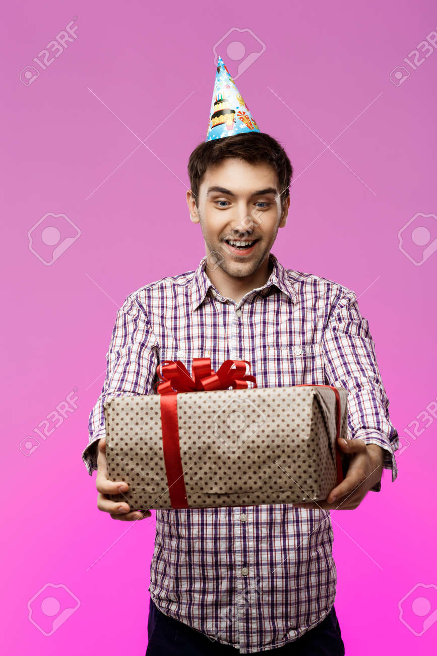 Happy Young Handsome Man Holding Birthday Gift In Box Over Purple