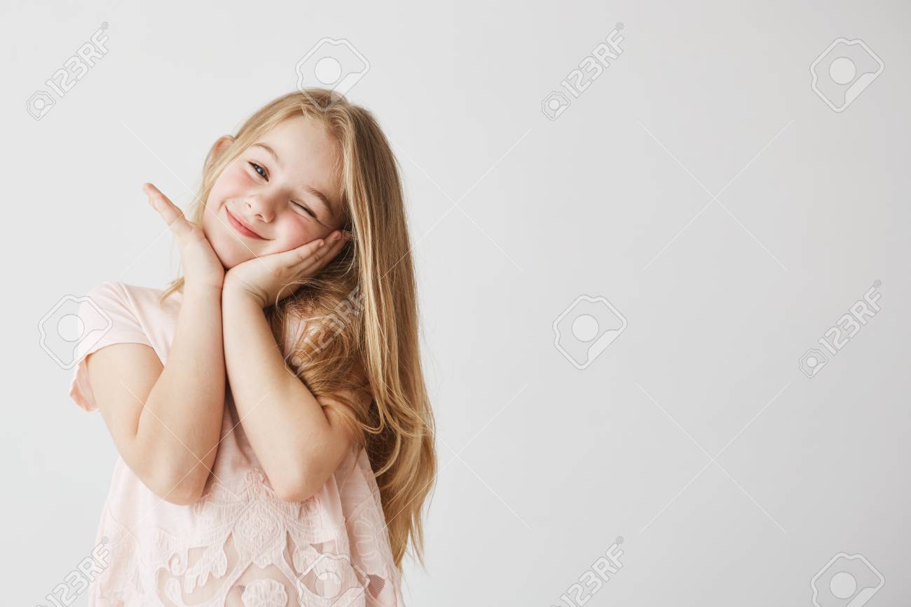 肌触り少しブロンドの女の子のポーズ ピンクのかわいいドレスで彼女の手で顔に触れるカメラのまばたきで笑顔します 幸せと喜びを探している子 領域をコピーします の写真素材 画像素材 Image