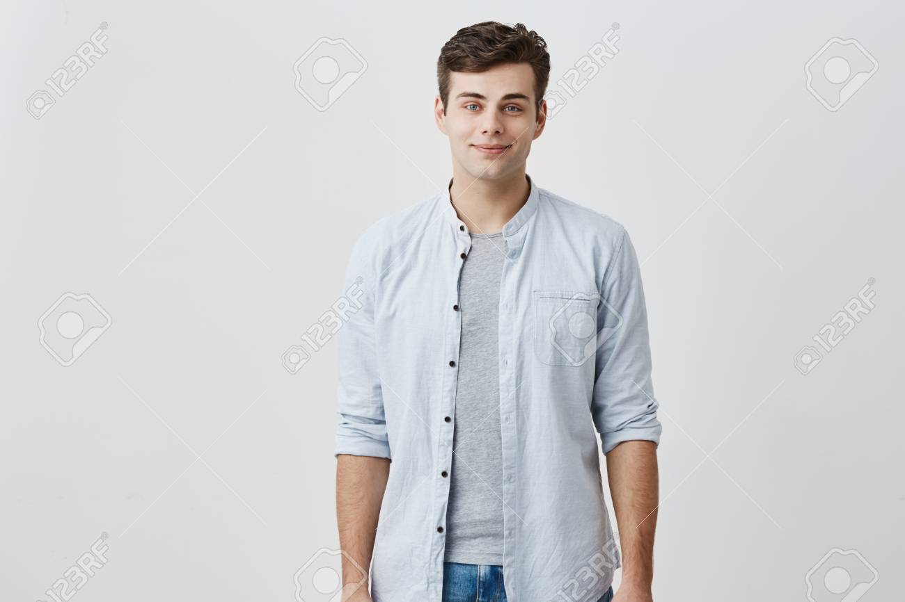 O Individuo Caucasiano Satisfeito Alegre Com Olhos Azuis E Cabelo Escuro Vestiu Se Na Camisa Azul Elegante Que Sorri No Bom Humor Ao Levantar No Estudio Contra O Fundo Cinzento Conceito Expressoes Faciais