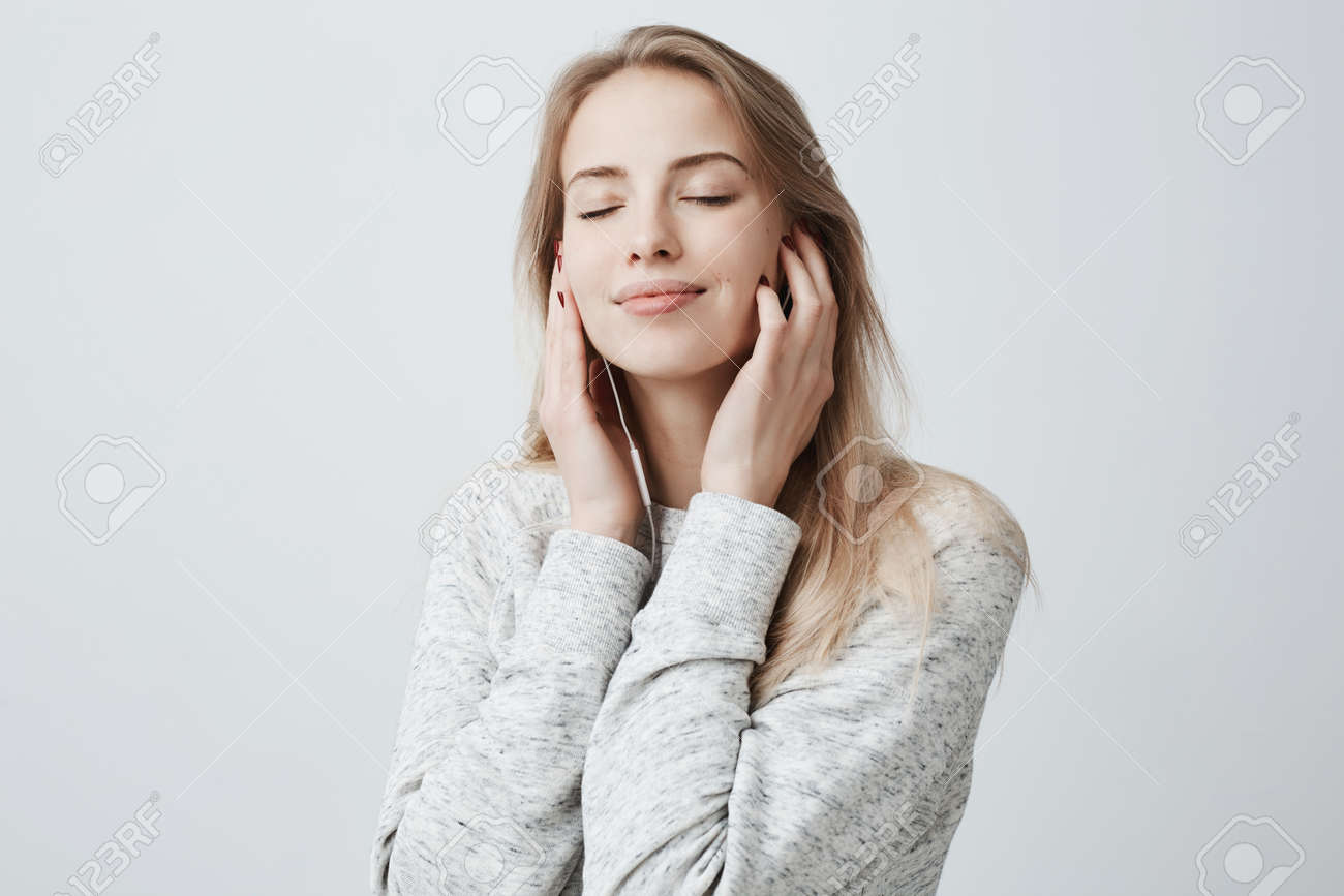 Close-up portrait of beautiful attractive European young woman in gray loose sweater, relaxing with closed eyes, listening to her favourite songs via white earphones, using music app. - 93438342 Close-up portrait of beautiful attractive European young woman in gray loose sweater, relaxing with closed eyes, listening to her favourite songs via white earphones, using music app. - 93438342