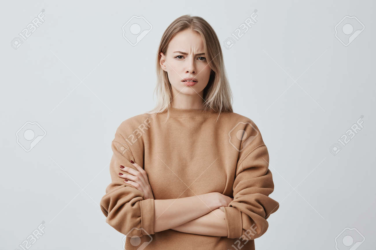 Waist-up portrait of beautiful girl with blonde straight hair frowning her face in displeasure, wearing loose long-sleeved sweater, keeping arms folded. Attractive young woman in closed posture. - 93873312 Waist-up portrait of beautiful girl with blonde straight hair frowning her face in displeasure, wearing loose long-sleeved sweater, keeping arms folded. Attractive young woman in closed posture. - 93873312
