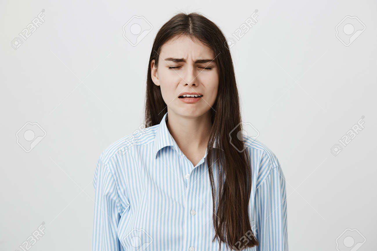 ブルーカラーのシャツを着て灰色の背景の上に立って 眉をひそめた眉毛と目を閉じて泣きそうになる悲しくて失望した大人の女性 疲れた事務員は彼女の友人に文句を言う の写真素材 画像素材 Image