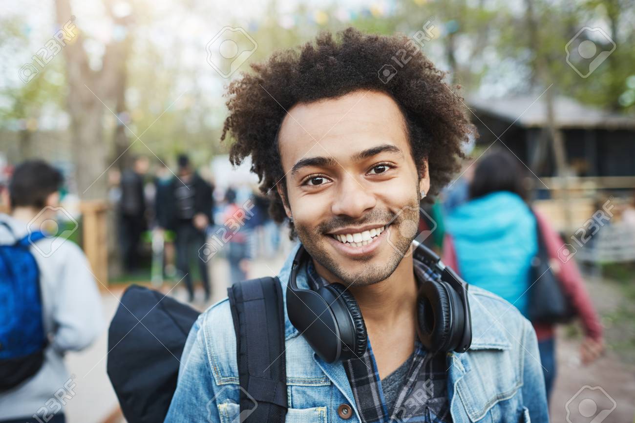 アフロの髪型と剛毛を持つ幸せな感情的な若いアフリカ系アメリカ人の男のクローズアップショットは デニムコートとバックパックを着用しながら広く微笑み フェスティバル中に公園を横断します の写真素材 画像素材 Image