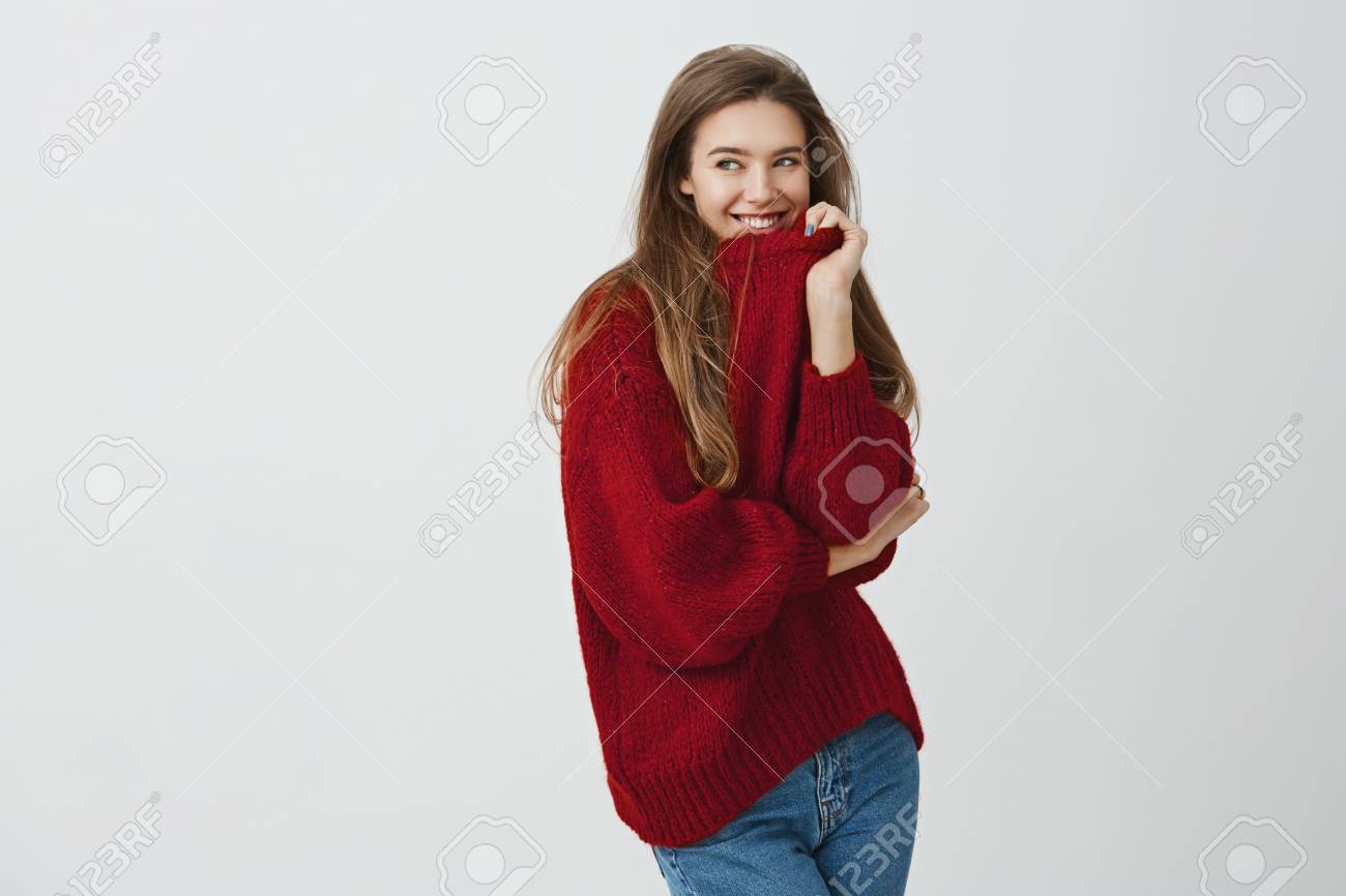 快適でゆるい服のような女の子 半ターン立ちながら襟を引っ張る赤いセーターを着た見栄えの良い魅力的な女性のスタジオショット 笑顔といちゃつく表情で脇を見て の写真素材 画像素材 Image
